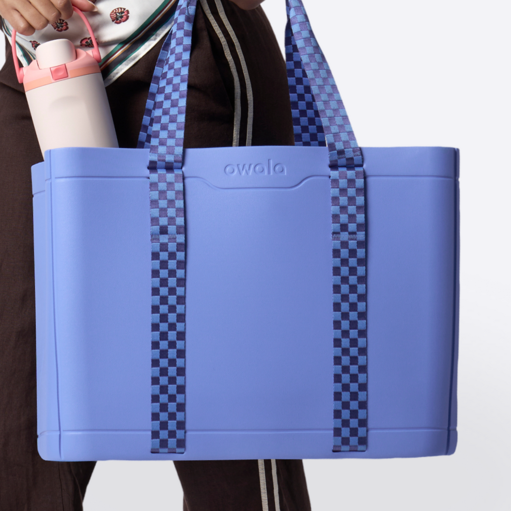 Model in yellow dress holding the Sun-Day Collapsible Tote in Wavelength, periwinkle blue body with blue checker straps and a small Owala bottle, against a blue draped backdrop